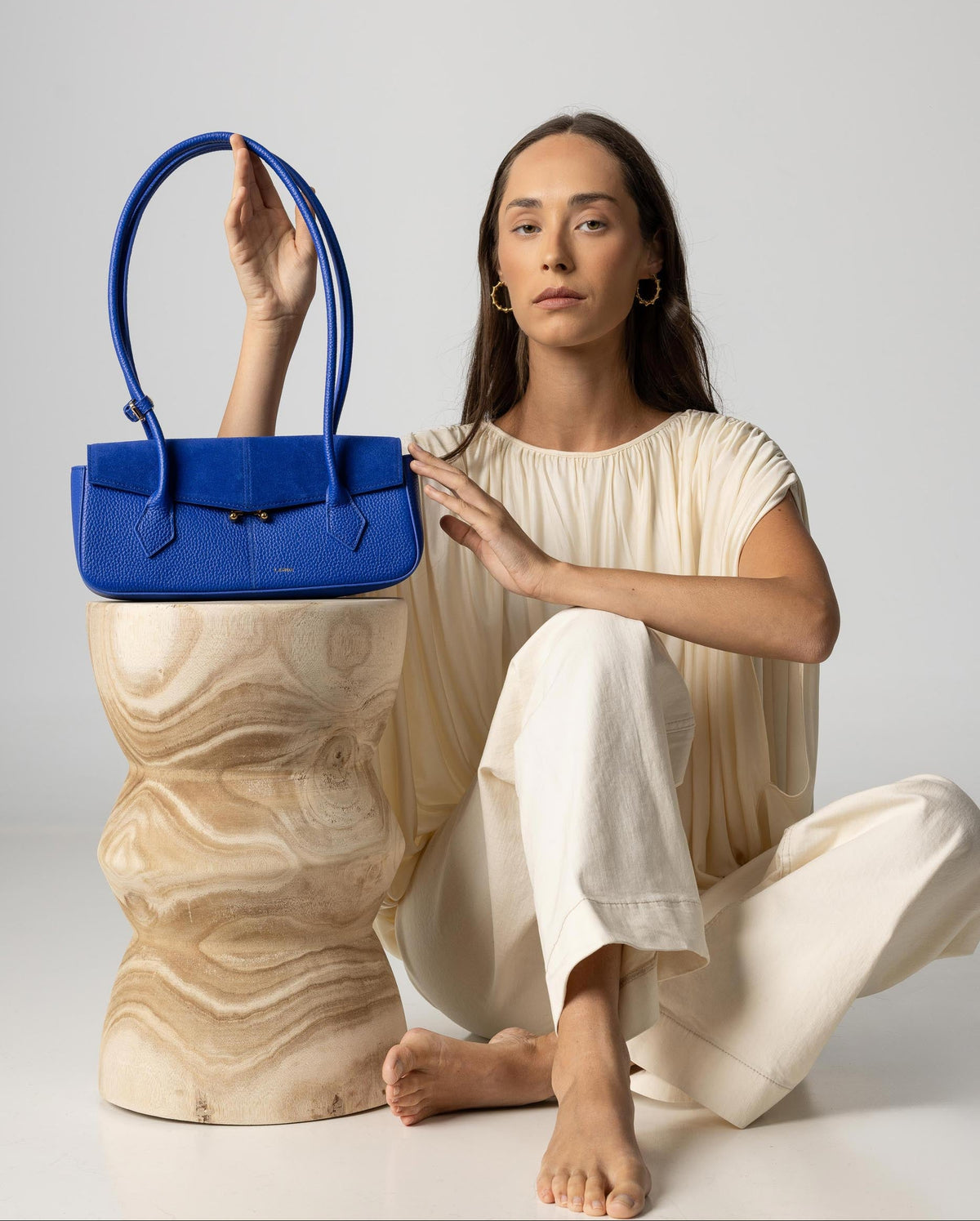 A woman in a cream-colored outfit sits on a sculptural wooden pedestal, holding a bright blue leather handbag. The background is a clean, neutral light gray studio backdrop.