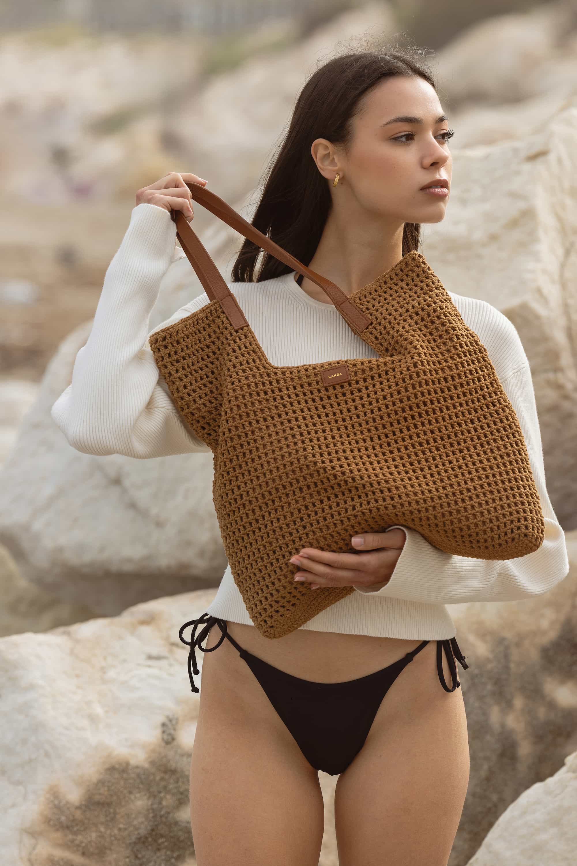 Woman holding a brown woven bag with tan leather handles on a rocky beach