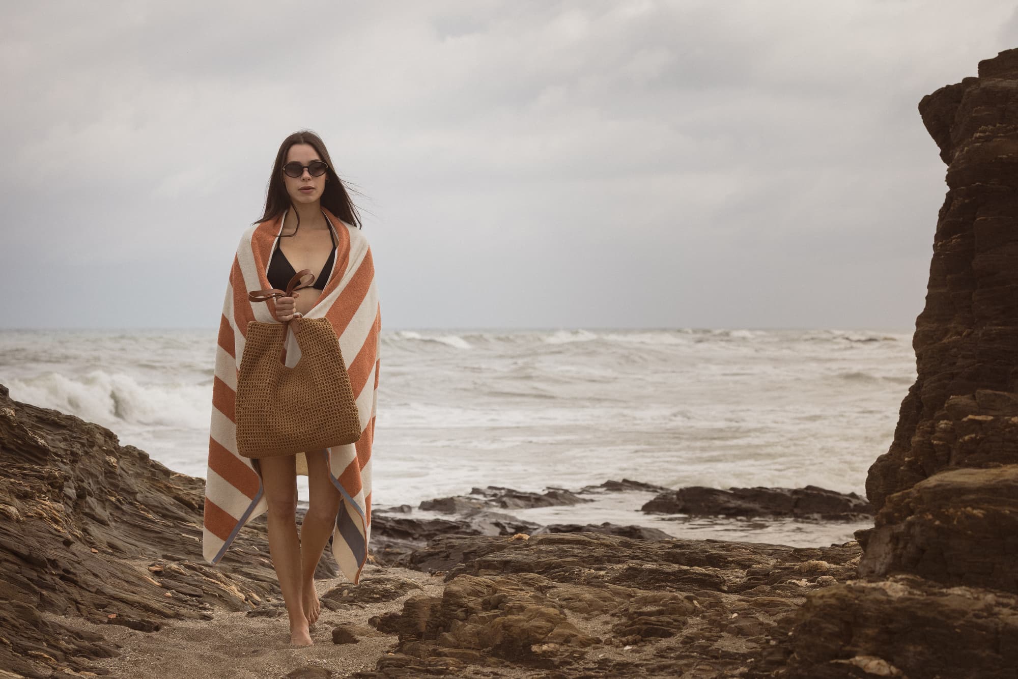 Woman wrapped in a striped towel holding a brown woven bag on a rocky beach with ocean waves in the background